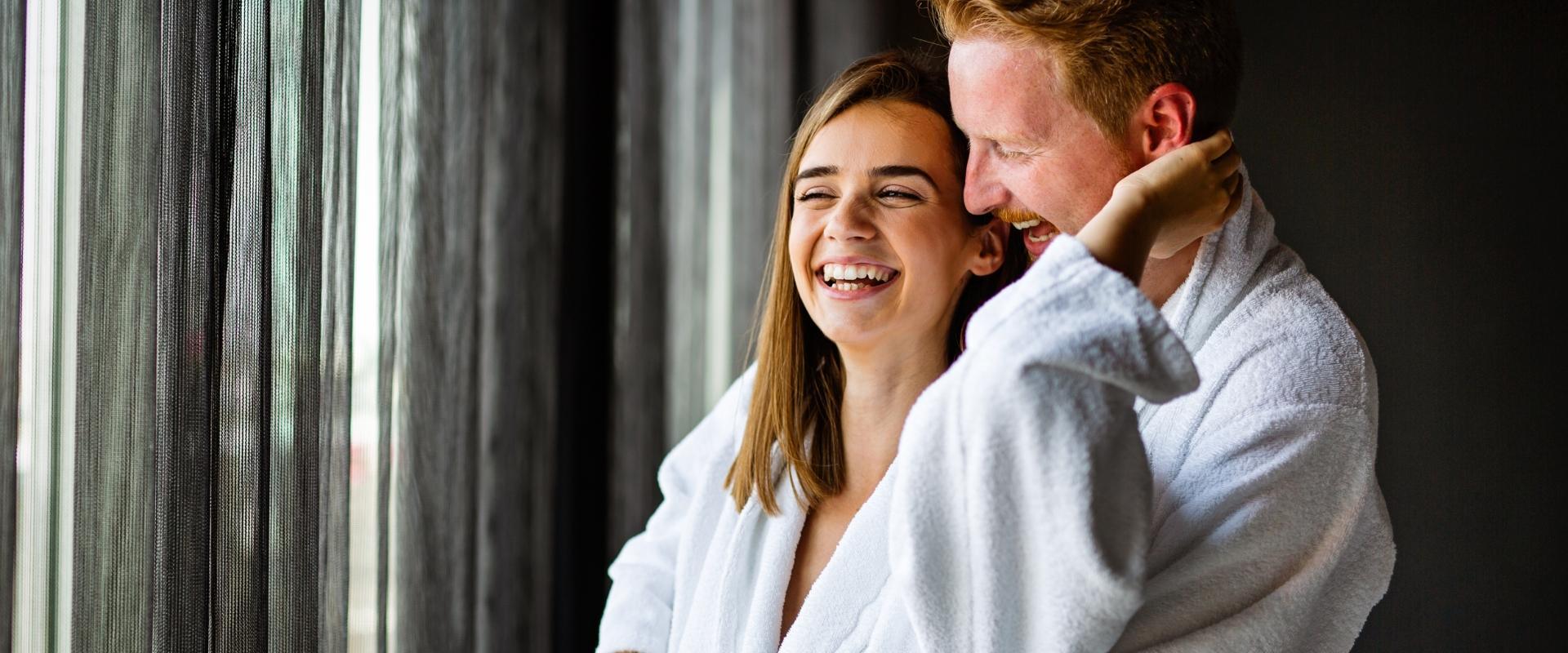 Coppia felice in accappatoi bianchi sorridenti vicino a una finestra