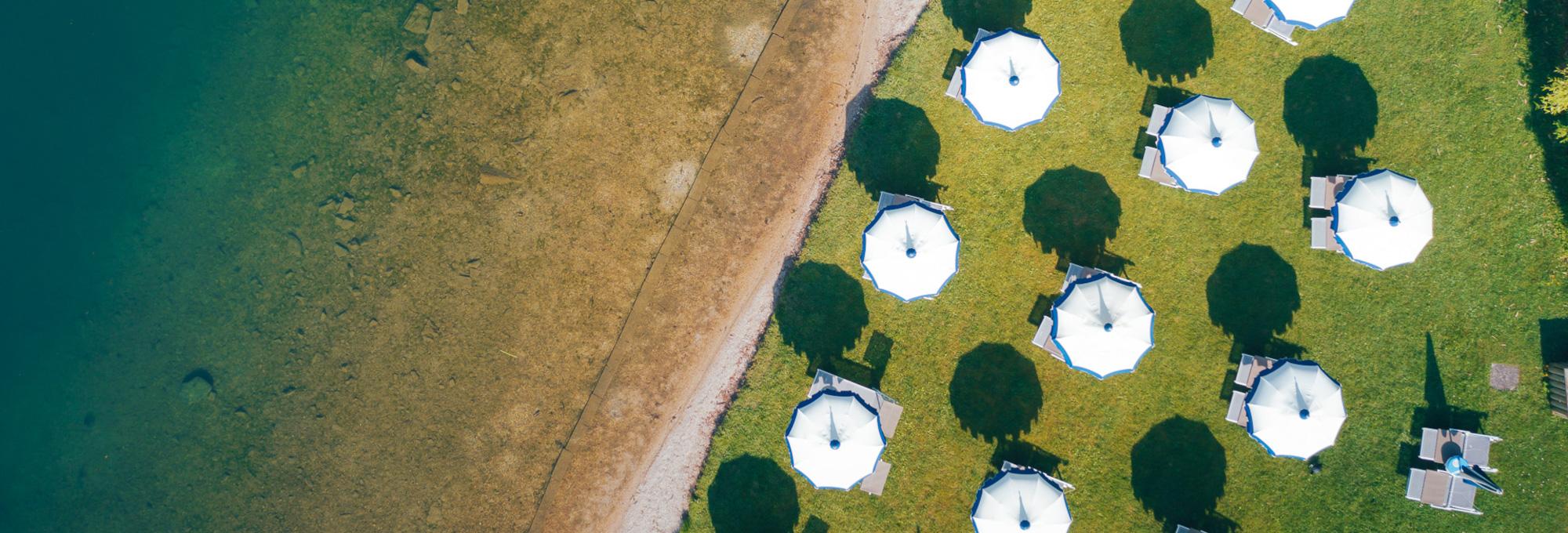 Vista aerea della spiaggia La Taverna con ombrelloni bianchi su prato affacciato al Lago di Levico