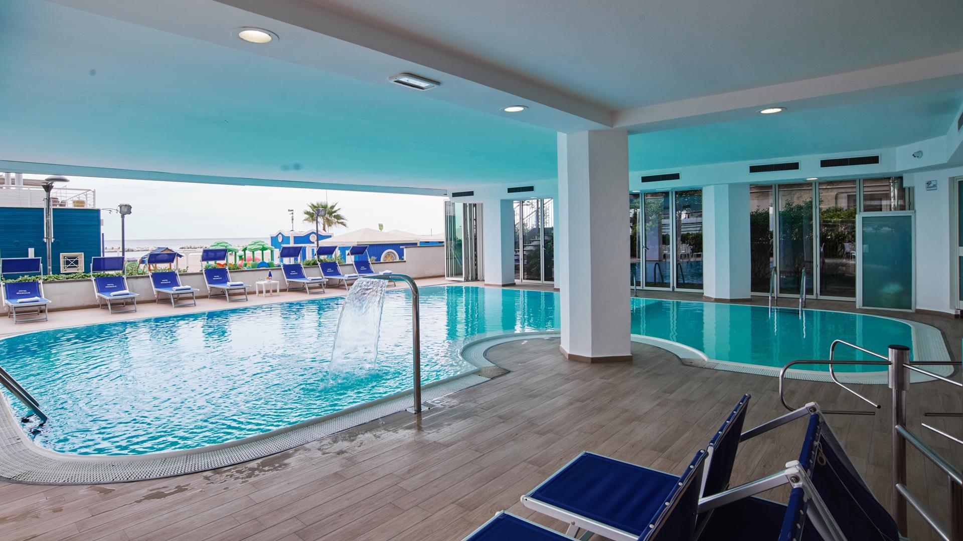 Piscina coperta con vista mare, lettini blu e cascata d'acqua